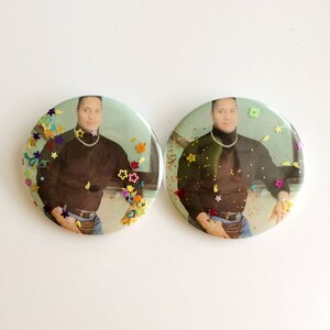 May include: Two round buttons with a photo of a person wearing a black turtleneck shirt and blue jeans. The buttons are covered in colorful glitter.