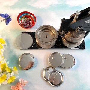 May include: A black button maker machine with a silver die and four silver button blanks. The machine is on a white surface with a small bowl of glitter and a butterfly decoration.