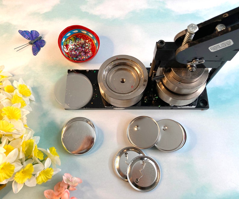 May include: A black button maker machine with a silver die and four silver button blanks. The machine is on a white surface with a small bowl of glitter and a butterfly decoration.