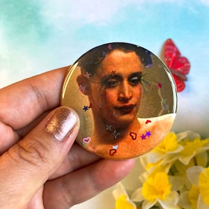 Puede incluir: Un botón redondo con un retrato de una persona de tez clara y cabello oscuro. La persona lleva una camisa blanca y tiene una expresión neutral. El botón está decorado con corazones de purpurina rosa y roja y estrellas de purpurina plateadas.