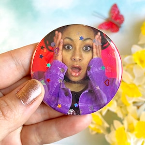 May include: A round button featuring a red background and a picture of a woman with her hands on her head, looking surprised. The button is covered in colourful glitter stars and a red heart.