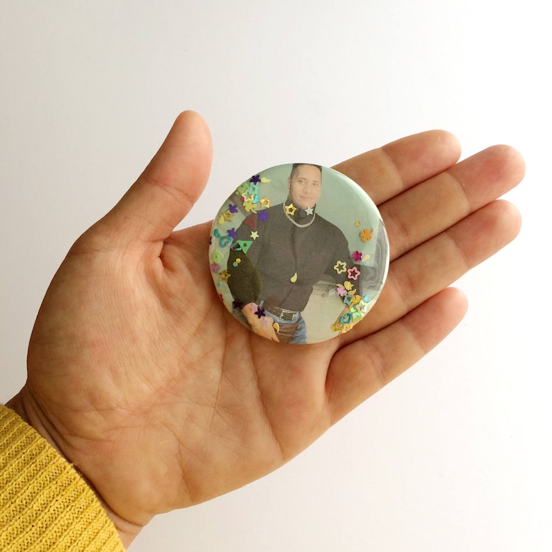 May include: A round button with a photo of a person wearing a black shirt and jeans. The button is covered in colorful glitter.
