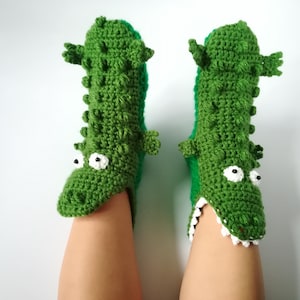 Könnte beinhalten: Ein Paar grüne Häkelslipper in Alligatorform. Die Slipper haben weiße Augen, eine Reihe weißer Zähne und kleine Arme. Die Slipper sind aus einem strukturierten grünen Garn gefertigt und bedecken Füße und Knöchel.