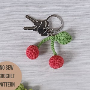 Puede incluir: Un llavero de cereza de crochet con un tallo y hojas verdes. El llavero está unido a un llavero plateado con dos llaves. El texto "NO SEW CROCHET PATTERN" es visible en la imagen.