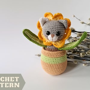 May include: A crocheted gray cat wearing a yellow flower pot with green leaves. The cat is sitting in a crocheted orange pot with green stripes.