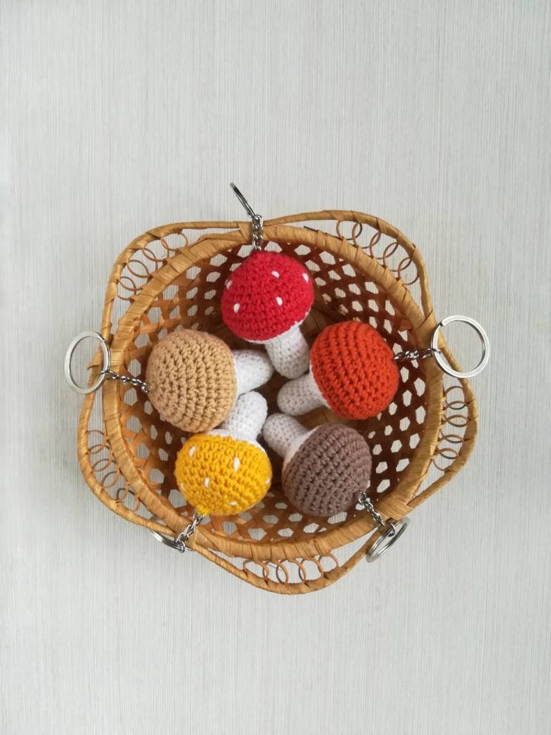 May include: Five crocheted mushroom keychains in a wicker basket. The mushrooms are red, orange, yellow, brown, and beige. Each mushroom has a white stem.