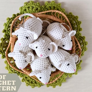 May include: A collection of white crochet sheep keychains with black eyes and pink noses, nestled in a woven basket atop a green crocheted doily. The image includes the text "PDF CROCHET PATTERN".