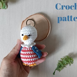 May include: Crochet pattern for a small amigurumi eagle keychain. The eagle is white with red and blue stripes and stars on its chest. It has a yellow beak and black eyes.