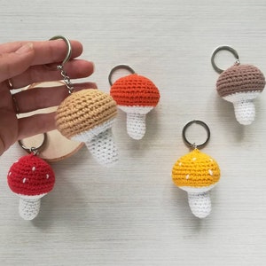 May include: Five crocheted mushroom keychains in different colors. The keychains are made of yarn and have a white stem and a colored cap. The caps are red, orange, brown, yellow, and beige.