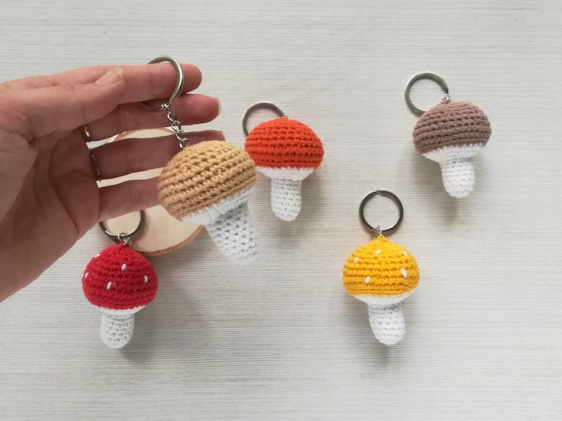 May include: Five crocheted mushroom keychains in different colors. The keychains are made of yarn and have a white stem and a colored cap. The caps are red, orange, brown, yellow, and beige.