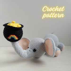 May include: A crocheted grey elephant toy holding a black pot of gold in its trunk. The pot has a rainbow and yellow gold. The text "Crochet pattern" is in yellow. The elephant has pink inner ears and feet.