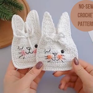 May include: Two white crocheted bunny-shaped pouches with embroidered eyes and pink noses. The pouches are held in the hands of a person. The text "NO-SEW CROCHET PATTERN" is visible in the upper right corner of the image.