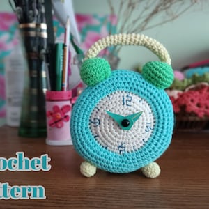 以下が含まれることがあります： 緑と青の顔と、黒の数字が書かれた白い時計の文字盤を持つ、かぎ針編みの目覚まし時計。時計は黄色のハンドルと緑と黄色の足を持っています。