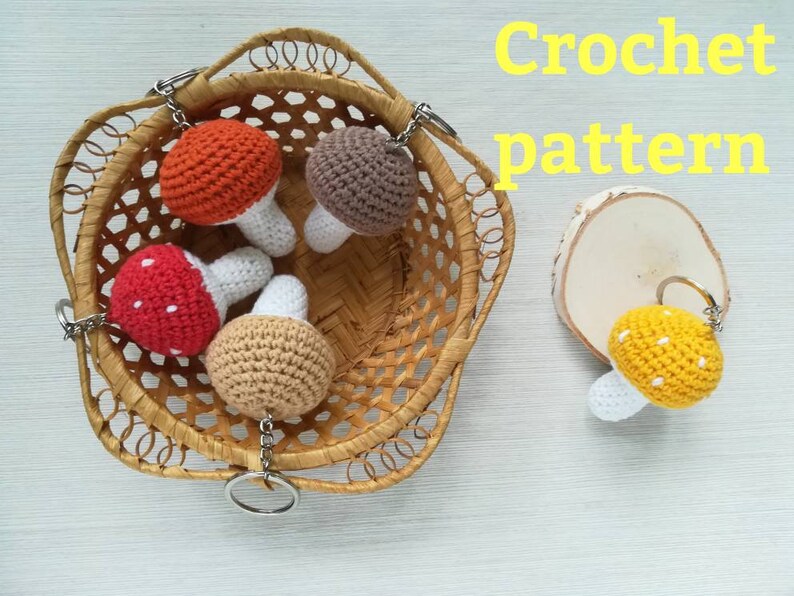 May include: Crochet pattern for mushroom keychains. The image shows four crocheted mushrooms with different colored caps: red, brown, beige, and yellow. Each mushroom has a white stem and a silver keychain attached.