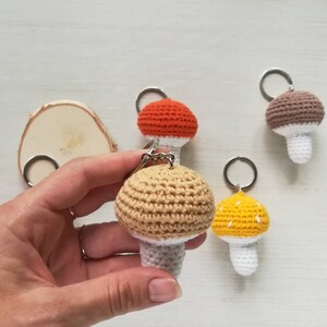 May include: Four crocheted mushroom keychains in different colors: brown, orange, yellow, and beige. Each mushroom has a white stem and a crocheted ring for attaching keys.