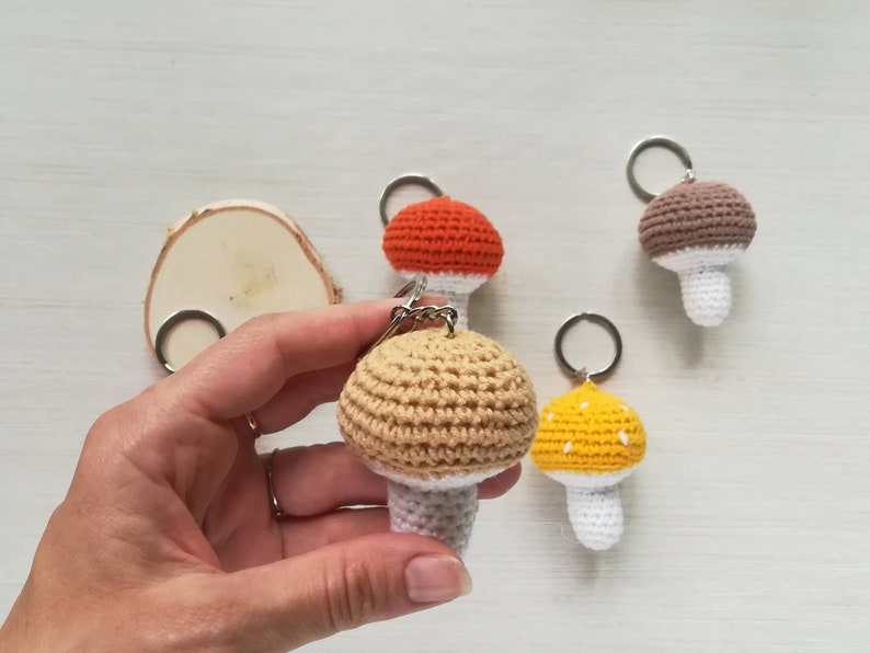 May include: Four crocheted mushroom keychains in different colors: brown, orange, yellow, and beige. Each mushroom has a white stem and a crocheted ring for attaching keys.