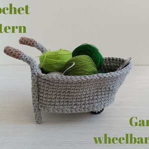 May include: A grey crocheted garden wheelbarrow filled with green and white balls of yarn. The text "Crochet pattern" is at the top of the image and "Garden wheelbarrow" is at the bottom.