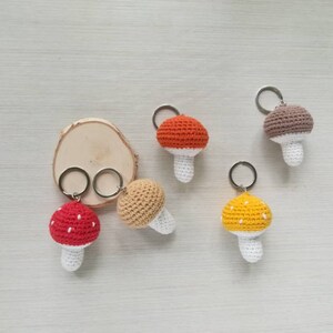 May include: Five crocheted mushroom keychains in different colors, including red, orange, yellow, and brown. Each mushroom has a white stem and a crocheted cap with white dots.