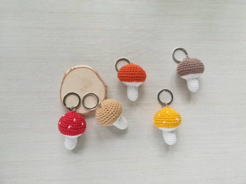 May include: Five crocheted mushroom keychains in different colors, including red, orange, yellow, and brown. Each mushroom has a white stem and a crocheted cap with white dots.