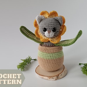 May include: Crochet pattern for a gray cat wearing a yellow sunflower and sitting in a green and brown pot. The cat has white ears and black eyes. The pot is on a wooden base. The text "CROCHET PATTERN" is at the bottom of the image.