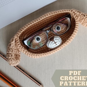 May include: A tan crochet eyeglass case shaped like a dog, holding a pair of eyeglasses. A small white crocheted detail is inside the case. The text "PDF CROCHET PATTERN" is in the lower right corner. Pencils are in the lower left.