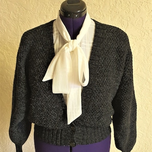 May include: A black knitted cardigan with a white bow tie. The cardigan has a v-neck and long sleeves.