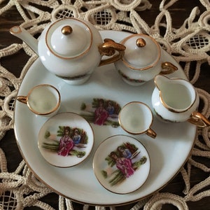 May include: A white porcelain tea set with gold accents. The set includes a teapot, creamer, sugar bowl, two teacups, and two saucers. Each piece features a delicate floral design with a couple in a romantic setting.