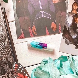 Może przedstawiać: Biały sztyft do ust z niebieskim i fioletowym wzorem. Na sztyfcie widnieje napis "Bookish Azriel".