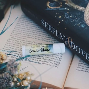 Könnte beinhalten: Ein weißer Lippenbalsam-Stift mit einem bunten Etikett, auf dem "Bookish Balms Lou le Blanc" steht. Der Lippenbalsam liegt auf einem geöffneten Buch mit dem sichtbaren Titel "Serpent & Dove".