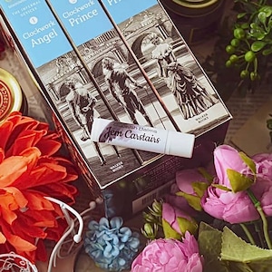 May include: Three hardcover books with illustrated covers, titled "Clockwork Angel", "Clockwork Prince", and "Clockwork Princess". A lip balm tube with the text "Brush Balms Jem Carstairs" is in the foreground.