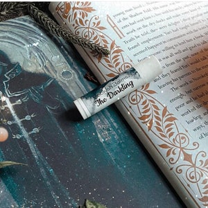 May include: A close-up of a white lip balm tube with black text that reads "Bookish Balms. The Darkling". The tube is lying on a book with a decorative gold border and text. The background is a dark blue and gray image of a person in a hooded cloak.
