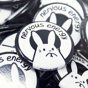 K&ouml;nnte beinhalten: Nahaufnahme eines Stapels schwarzer und wei&szlig;er Anstecknadeln mit einem wei&szlig;en Kaninchen und dem Text "nervous energy" auf jeder Nadel.