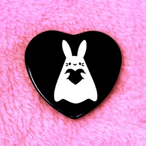 Puede incluir: Un pin negro en forma de corazón con un conejito de dibujos animados blanco sosteniendo un corazón. El conejito tiene orejas largas y una cara sonriente. El pin está sobre un fondo rosa y difuso.