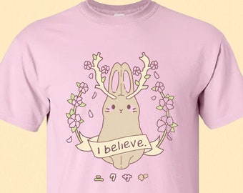 May include: Pink t-shirt with a brown bunny wearing antlers and a banner that says "I believe." The bunny is surrounded by pink flowers.
