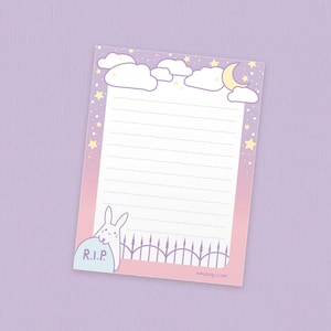 May include: A pink and purple lined notepad with a white rabbit peeking out from behind a tombstone that says "R.I.P." The notepad has a night sky border with clouds, stars, and a crescent moon.