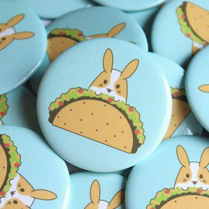 May include: A collection of blue and white round buttons with a cartoon image of a bunny rabbit peeking out of a taco. The taco is filled with lettuce, tomatoes, and cheese.