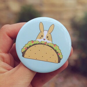 May include: A blue button with a cartoon image of a bunny rabbit inside a taco. The taco is filled with lettuce, tomato, and cheese.