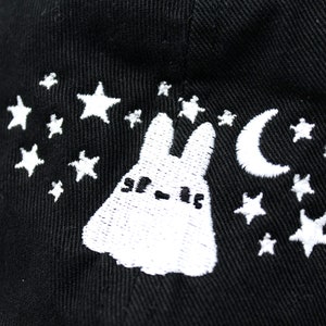 May include: Black baseball cap with white embroidered design featuring a cartoon bunny, crescent moon, and stars. The cap has a curved brim and a structured crown. The design is a whimsical, night-themed illustration.