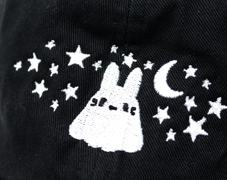 May include: Black baseball cap with white embroidered design featuring a cartoon bunny, crescent moon, and stars. The cap has a curved brim and a structured crown. The design is a whimsical, night-themed illustration.