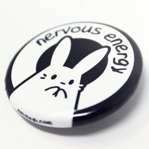 K&ouml;nnte beinhalten: Ein schwarz-wei&szlig;er Knopf mit einem wei&szlig;en Kaninchen mit schwarzem Umriss und dem Text "nervous energy" in einem schwarzen Kreis.