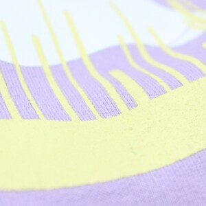 May include: Close-up of a lavender colored fabric with a yellow graphic design. The design is a series of vertical lines that are slightly curved.