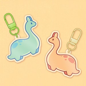 May include: Two dinosaur-shaped keychains. One is light blue with darker blue spots and a green clasp. The other is peach with reddish-brown spots and a gold clasp. Both have a cartoon style and are on a light yellow background.