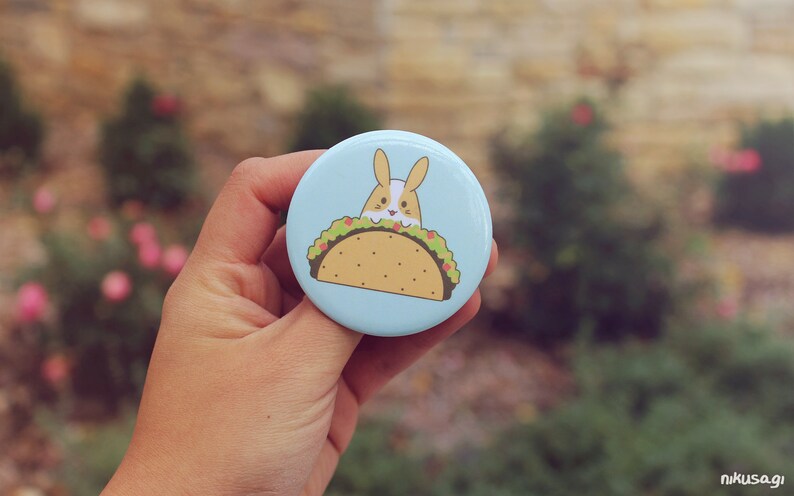 May include: A blue button with a cartoon image of a bunny rabbit inside a taco. The taco is filled with lettuce, tomato, and cheese.