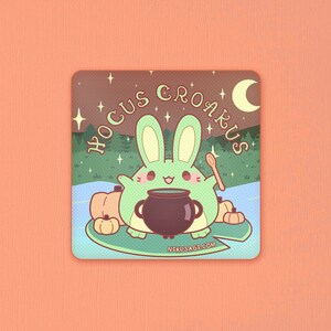 May include: A green bunny rabbit with a cauldron and a wooden spoon, surrounded by pumpkins. The text "Hocus Croakus" is written in a whimsical font above the bunny. The background is a dark brown with stars and a crescent moon.