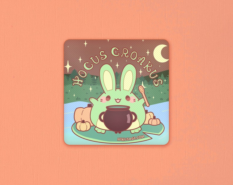 May include: A green bunny rabbit with a cauldron and a wooden spoon, surrounded by pumpkins. The text "Hocus Croakus" is written in a whimsical font above the bunny. The background is a dark brown with stars and a crescent moon.