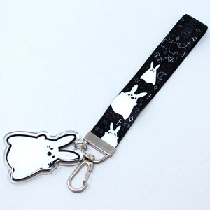 May include: A keychain with a white bunny charm and a black lanyard. The lanyard displays a pattern of white bunnies, bats, moons, stars, and crosses. The keychain is connected with a silver-coloured clasp.