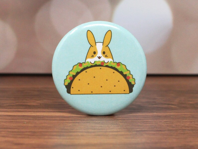May include: A light blue button with a cartoon image of a bunny rabbit wearing a taco hat. The taco is filled with lettuce, tomato, and cheese.