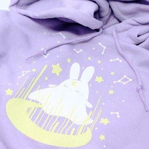 May include: A lavender hoodie with a white bunny wearing a crescent moon on its head. The bunny is surrounded by yellow stars and constellations.