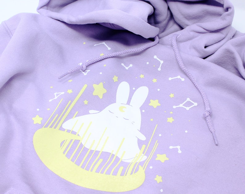 May include: A lavender hoodie with a white bunny wearing a crescent moon on its head. The bunny is surrounded by yellow stars and constellations.