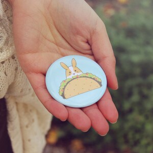 May include: A light blue button with a cartoon bunny rabbit wearing a taco. The taco is filled with lettuce, cheese, and meat.